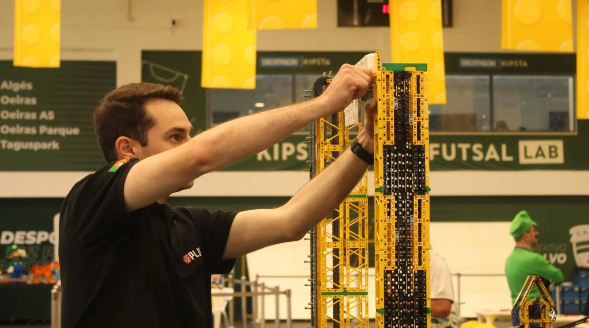 Person assembling large yellow and black LEGO structure at BRInCKa 2026 event in futsal facility.