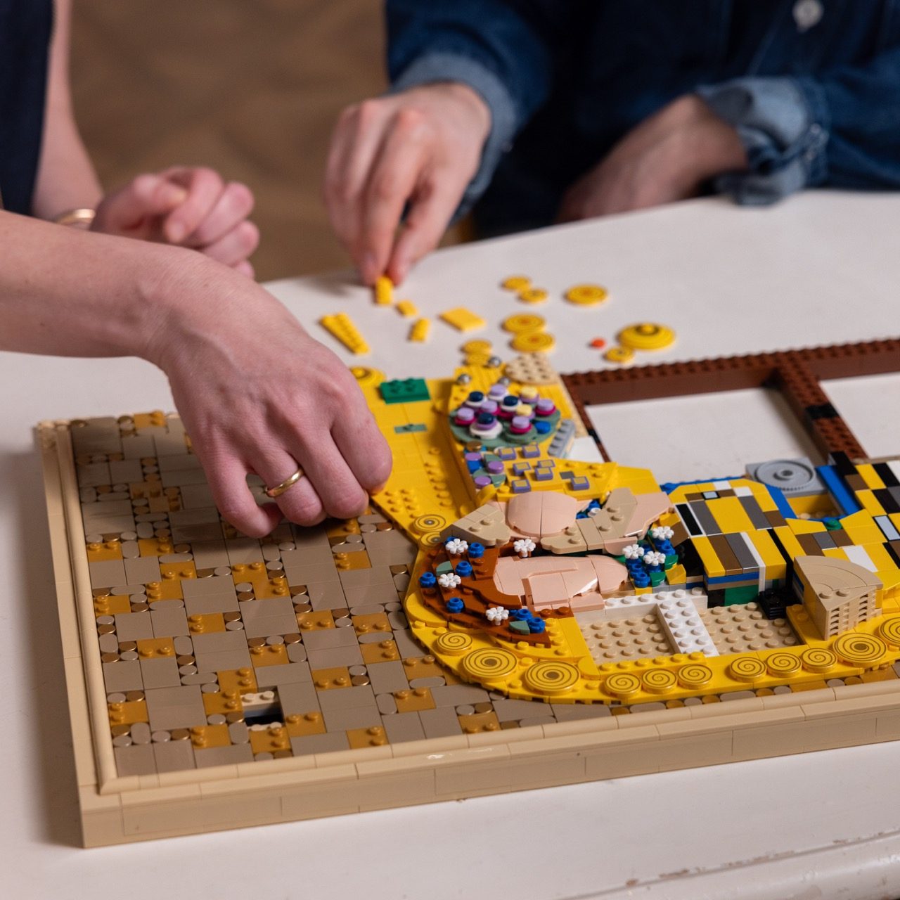 Hands assembling LEGO Art Gustav Klimt set with golden tiles and colorful decorative elements on wooden frame.