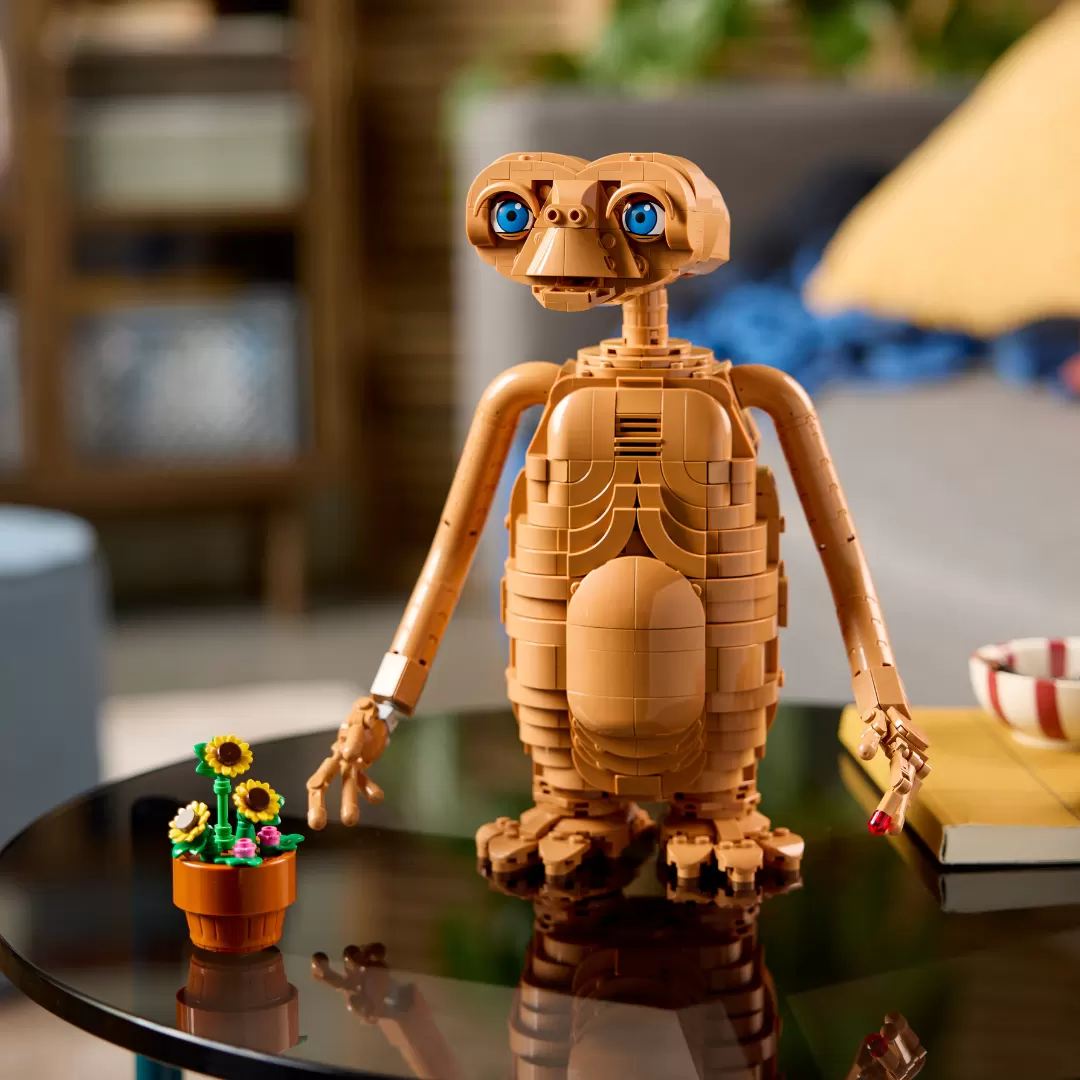 LEGO E.T. figure with glowing blue eyes and illuminated chest standing on black table next to potted flowers.