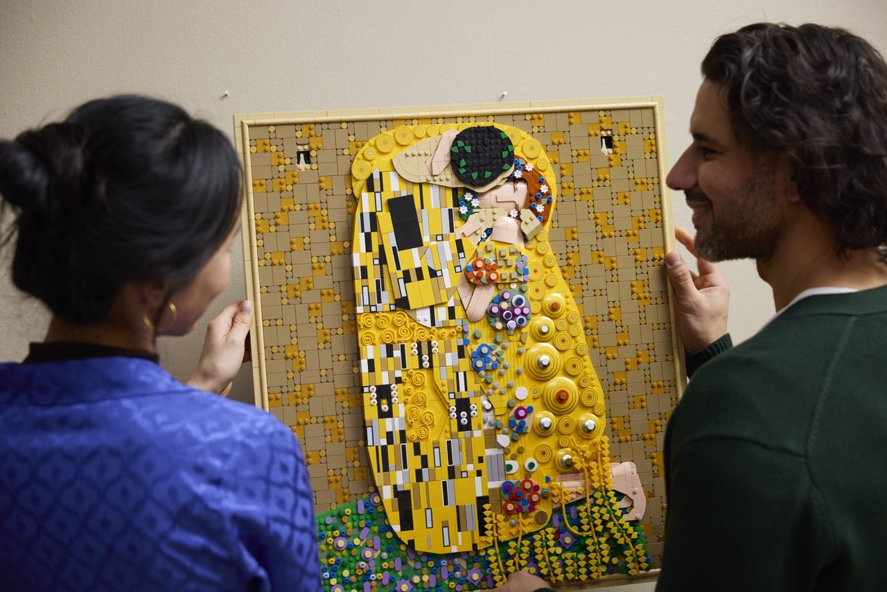 Two people admiring a large LEGO Art mosaic of Klimt's "The Kiss" with yellow, black, and colorful decorative elements.