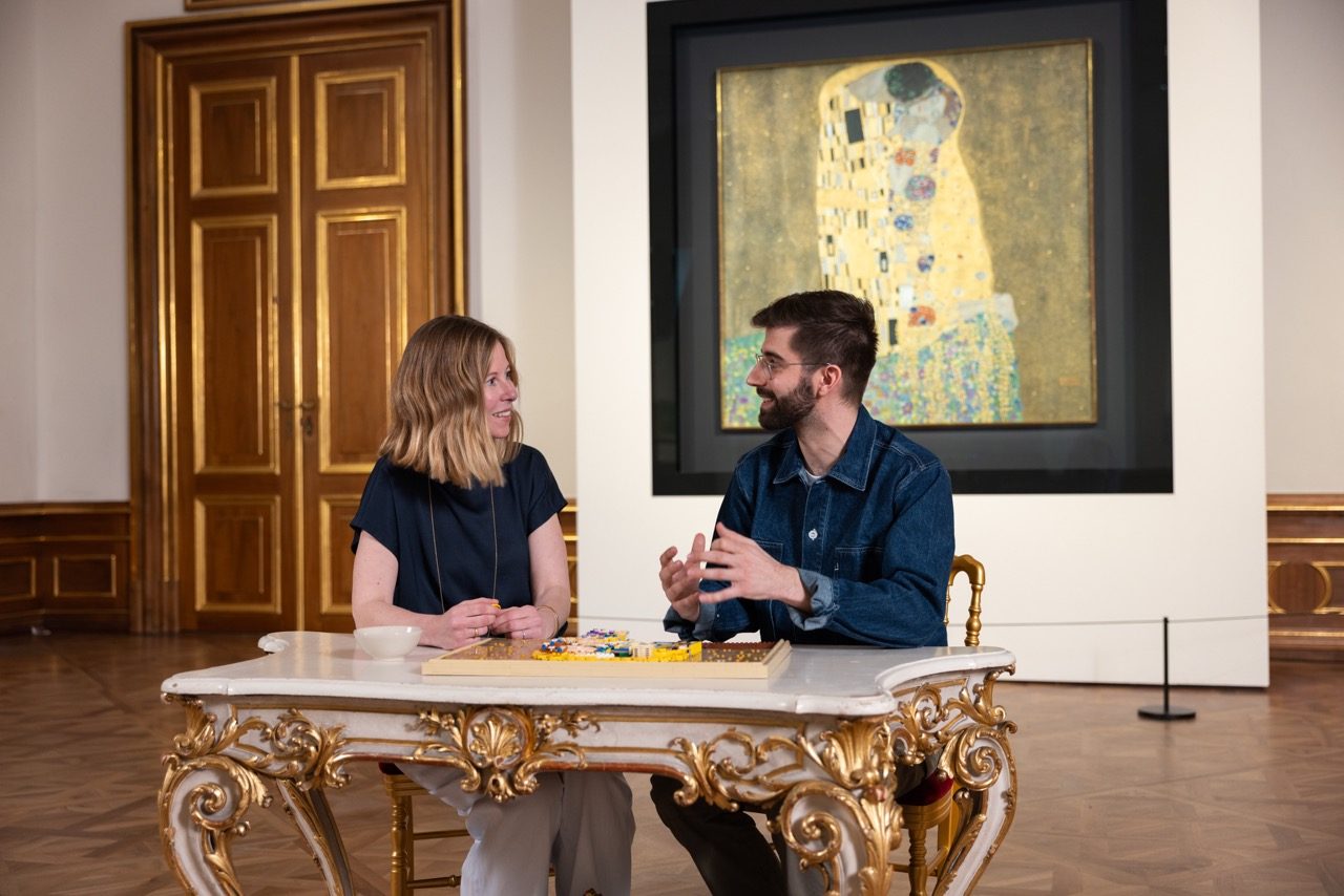 Two people examine LEGO Art Gustav Klimt set at ornate table with painting replica displayed behind.
