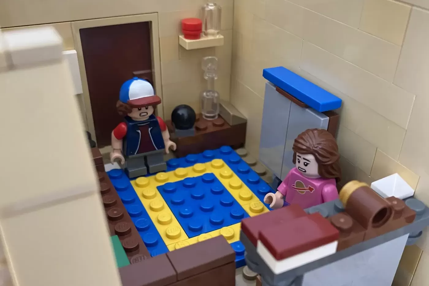 LEGO custom build of Mystery Shack bedroom with two minifigures, blue and yellow rug, bed, and bathroom fixtures.